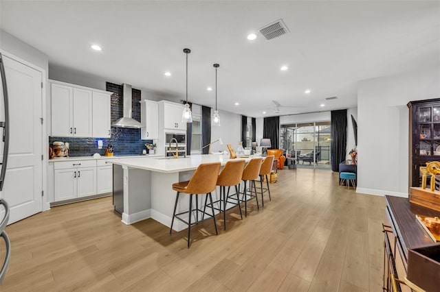 kitchen featuring wall chimney exhaust hood, light hardwood / wood-style flooring, hanging light fixtures, a center island with sink, and white cabinets