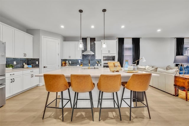 kitchen featuring built in microwave, white cabinetry, pendant lighting, a kitchen island with sink, and wall chimney range hood