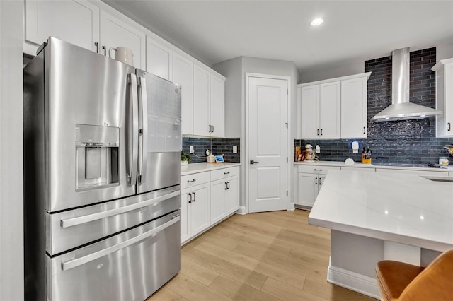 kitchen with white cabinets, decorative backsplash, stainless steel refrigerator with ice dispenser, wall chimney range hood, and light hardwood / wood-style flooring