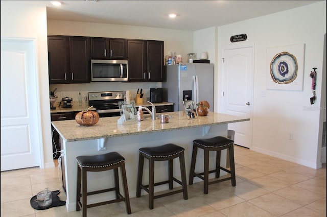 kitchen with dark brown cabinetry, appliances with stainless steel finishes, a center island with sink, and a kitchen bar