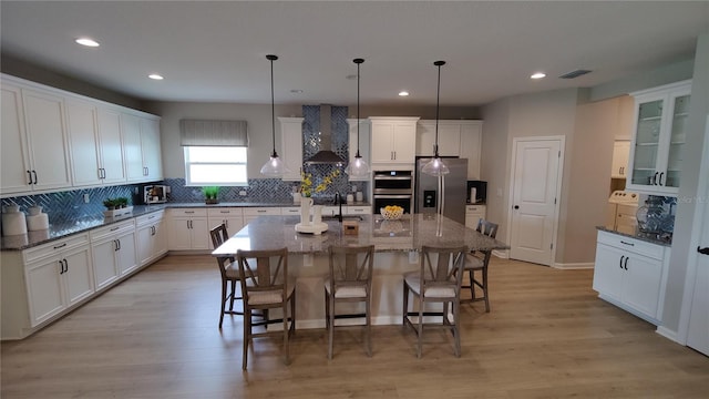 kitchen featuring wall chimney exhaust hood, a kitchen island with sink, pendant lighting, stainless steel appliances, and light hardwood / wood-style floors