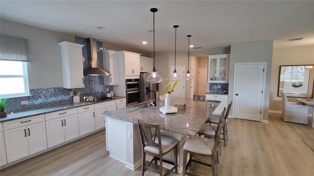 kitchen with wall chimney exhaust hood, sink, hanging light fixtures, dark stone countertops, and a kitchen island with sink