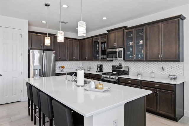 kitchen featuring decorative light fixtures, dark brown cabinets, appliances with stainless steel finishes, a kitchen island with sink, and decorative backsplash