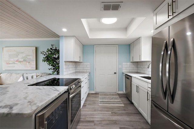 kitchen with white cabinets, backsplash, a tray ceiling, stainless steel appliances, and light hardwood / wood-style flooring