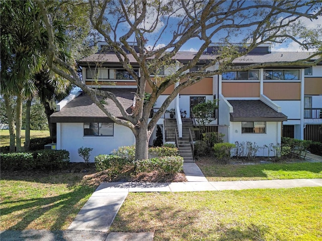 multi unit property with stucco siding, a shingled roof, stairs, and a front yard