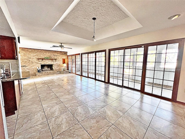 unfurnished living room featuring light tile patterned floors, ceiling fan, a tray ceiling, a brick fireplace, and a textured ceiling