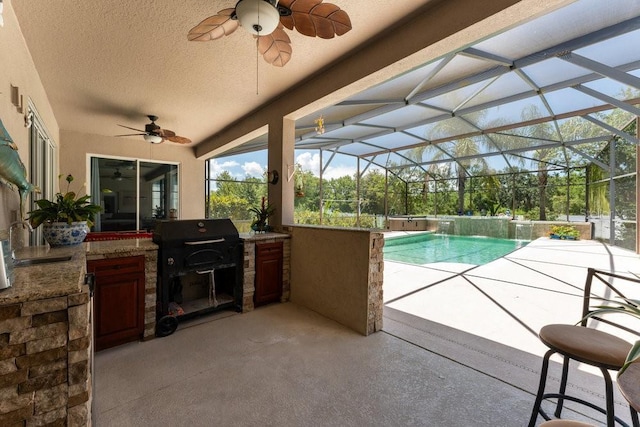 outdoor pool with area for grilling, a patio area, glass enclosure, ceiling fan, and exterior kitchen