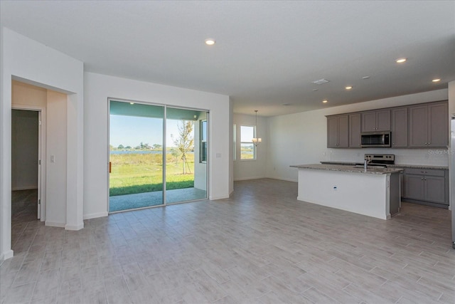 kitchen featuring appliances with stainless steel finishes, decorative light fixtures, an island with sink, light stone counters, and light wood-type flooring