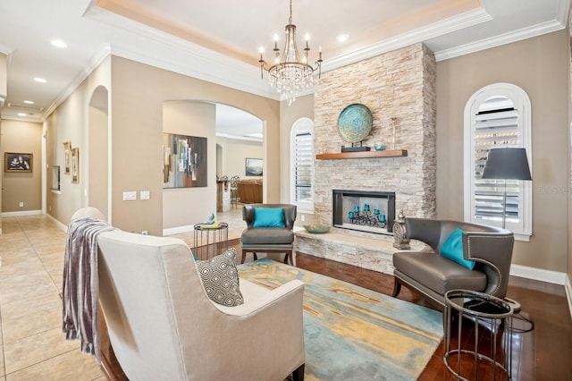 living room featuring light tile patterned flooring, a stone fireplace, crown molding, a raised ceiling, and a notable chandelier