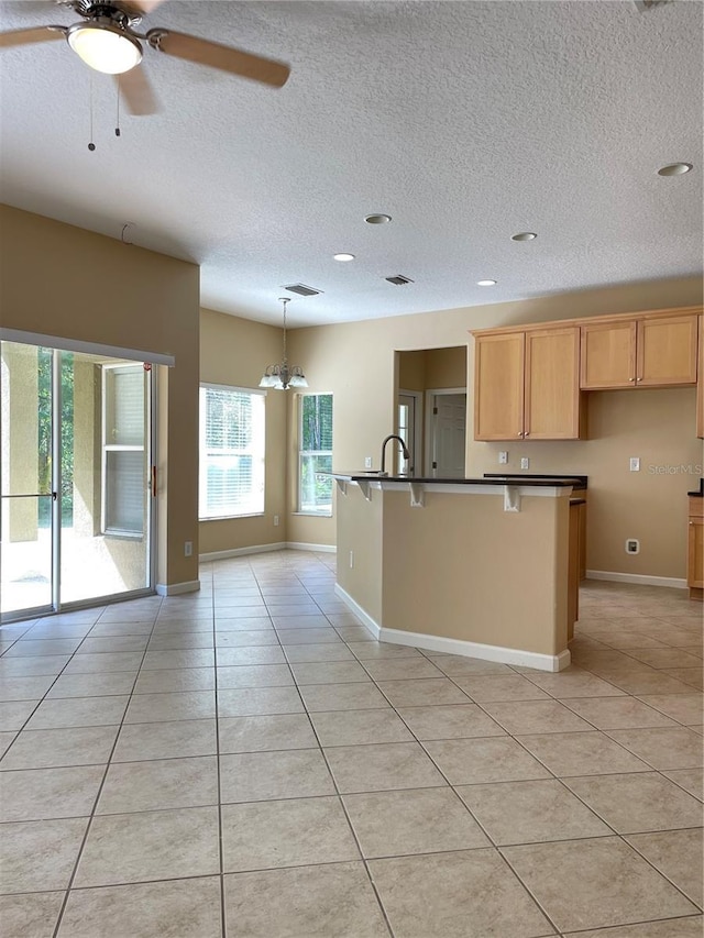 kitchen with light tile patterned floors, a textured ceiling, dark countertops, a kitchen island with sink, and ceiling fan with notable chandelier