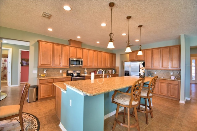 kitchen featuring appliances with stainless steel finishes, decorative light fixtures, light tile patterned floors, light stone counters, and a center island with sink