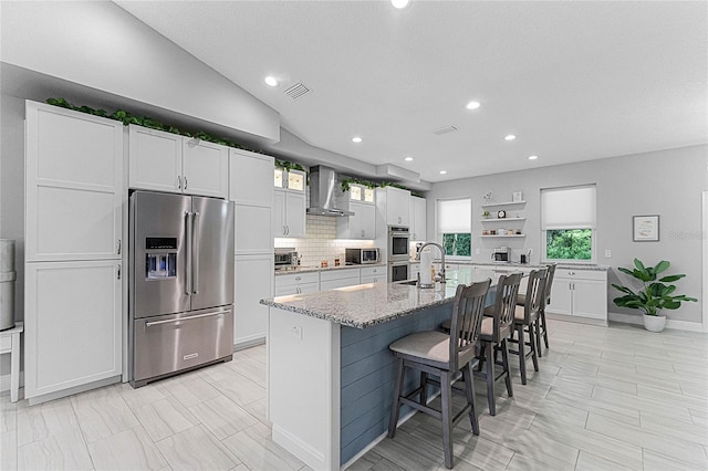 kitchen featuring wall chimney exhaust hood, appliances with stainless steel finishes, a kitchen breakfast bar, an island with sink, and white cabinets