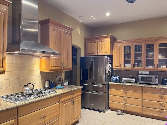 kitchen with light stone counters, stainless steel appliances, decorative backsplash, glass insert cabinets, and wall chimney exhaust hood