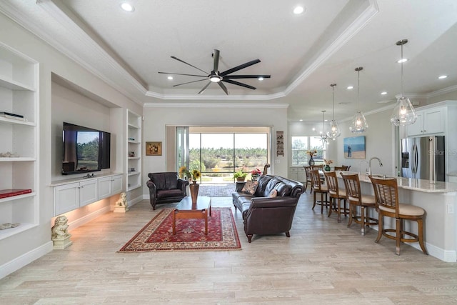 living room featuring crown molding, a tray ceiling, built in shelves, ceiling fan with notable chandelier, and light wood-type flooring