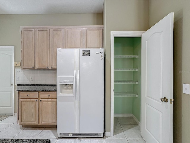 kitchen with dark stone countertops, white refrigerator with ice dispenser, light tile patterned flooring, light brown cabinetry, and decorative backsplash