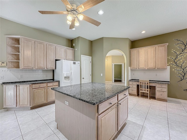 kitchen with light brown cabinetry, white fridge with ice dispenser, dark stone countertops, and a kitchen island