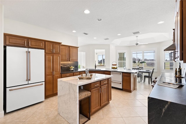 kitchen featuring light tile patterned floors, a kitchen island with sink, tasteful backsplash, high end white fridge, and dark stone counters