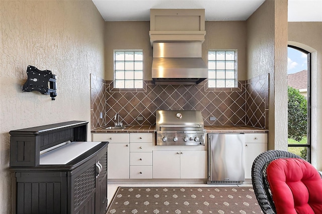 kitchen featuring freestanding refrigerator, a sink, wall chimney range hood, white cabinetry, and a wealth of natural light