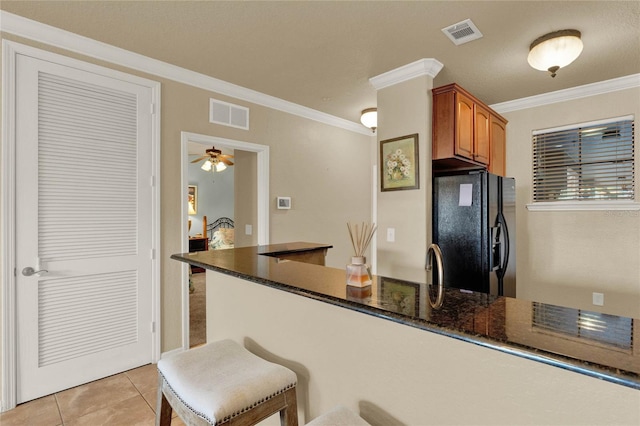 kitchen with light tile patterned floors, dark stone countertops, ornamental molding, a kitchen bar, and black fridge