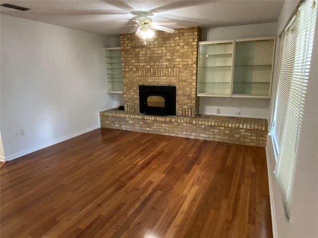 unfurnished living room with a brick fireplace, dark wood finished floors, visible vents, and a ceiling fan