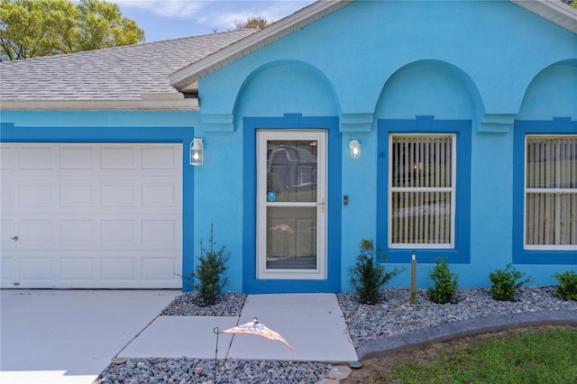 view of exterior entry with a garage, roof with shingles, and stucco siding