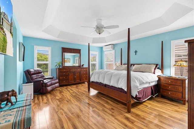 bedroom featuring light wood finished floors, ceiling fan, a tray ceiling, and a wall unit AC