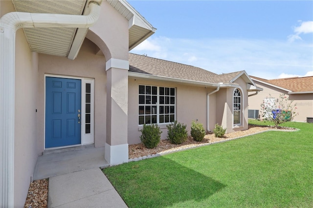 entrance to property featuring stucco siding, central AC unit, a shingled roof, and a lawn