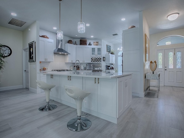 kitchen featuring a breakfast bar area, white cabinetry, light countertops, wall chimney range hood, and a center island with sink