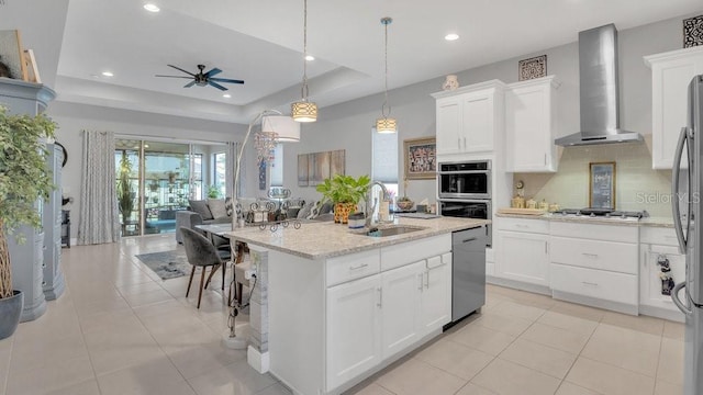 kitchen featuring stainless steel appliances, a sink, open floor plan, wall chimney exhaust hood, and a raised ceiling