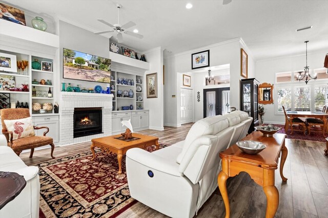 living room with hardwood / wood-style flooring, a brick fireplace, built in shelves, ornamental molding, and ceiling fan with notable chandelier