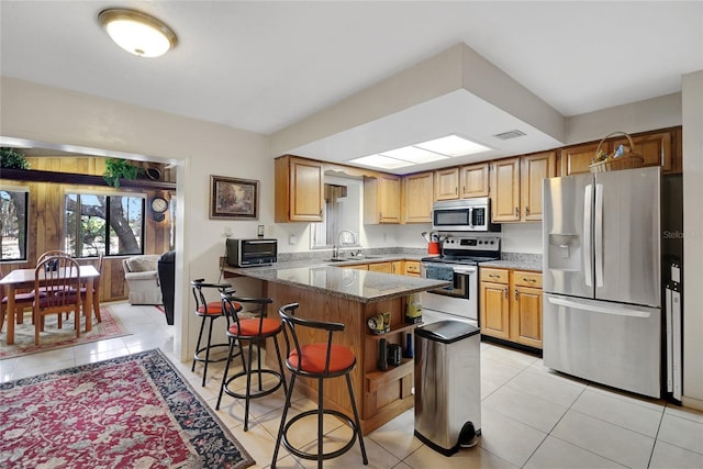 kitchen with light tile patterned floors, stainless steel appliances, a peninsula, a kitchen breakfast bar, and dark stone counters