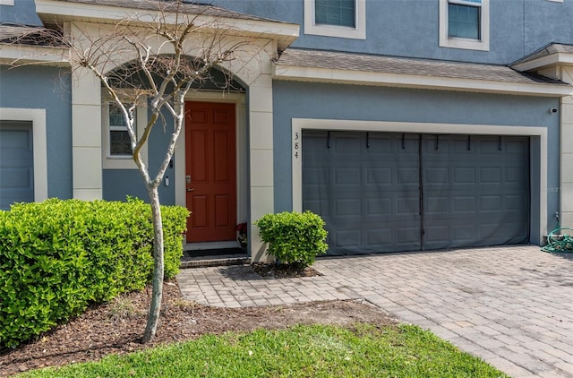 doorway to property featuring a garage, a shingled roof, decorative driveway, and stucco siding