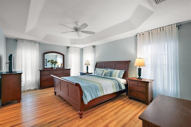 bedroom featuring a tray ceiling, multiple windows, ceiling fan, and light wood-style flooring