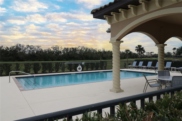 pool at dusk featuring a patio area, fence, and a community pool