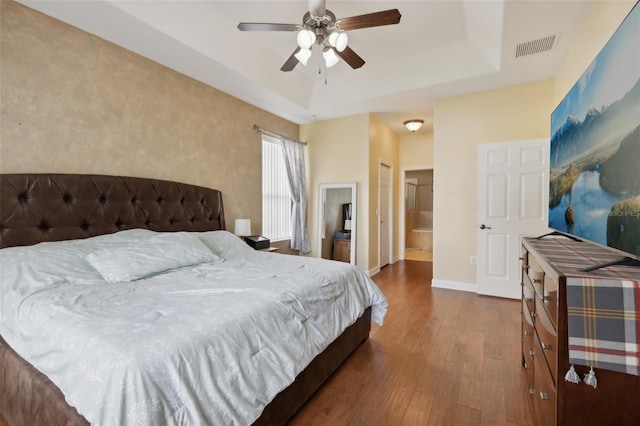 bedroom featuring baseboards, visible vents, a raised ceiling, ensuite bath, and wood finished floors