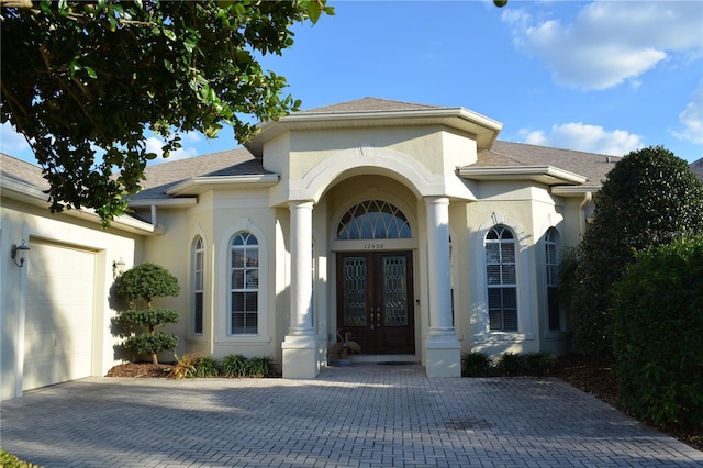 view of exterior entry featuring stucco siding, decorative driveway, french doors, roof with shingles, and an attached garage