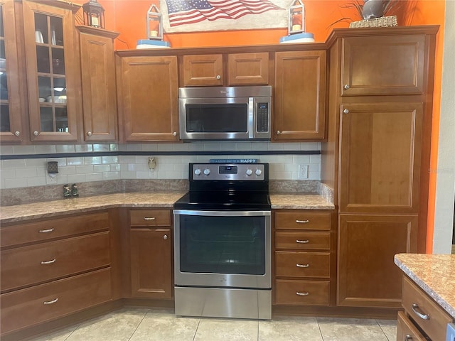 kitchen with tasteful backsplash, brown cabinetry, glass insert cabinets, light stone countertops, and stainless steel appliances