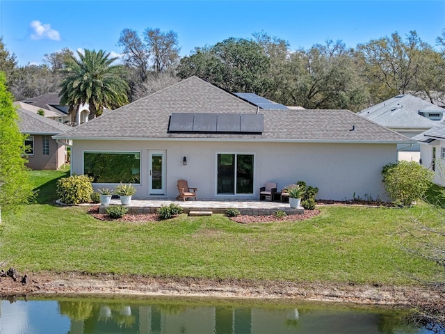 back of property featuring a shingled roof, a lawn, a water view, a patio area, and roof mounted solar panels