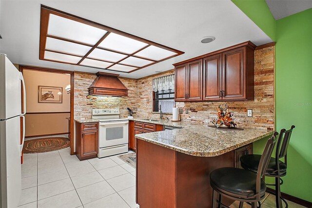 kitchen with light stone counters, custom exhaust hood, a sink, white appliances, and a peninsula