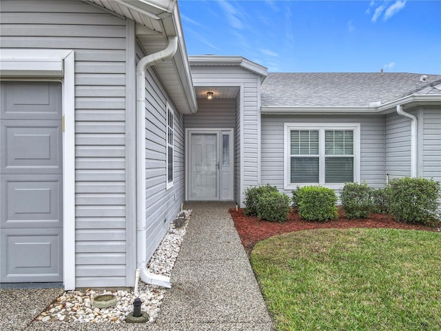 doorway to property featuring an attached garage, a lawn, and roof with shingles