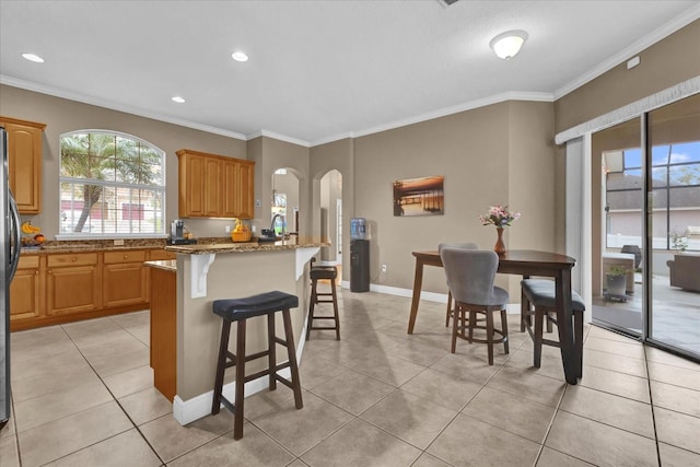 kitchen with arched walkways, light tile patterned floors, stone countertops, a kitchen breakfast bar, and crown molding