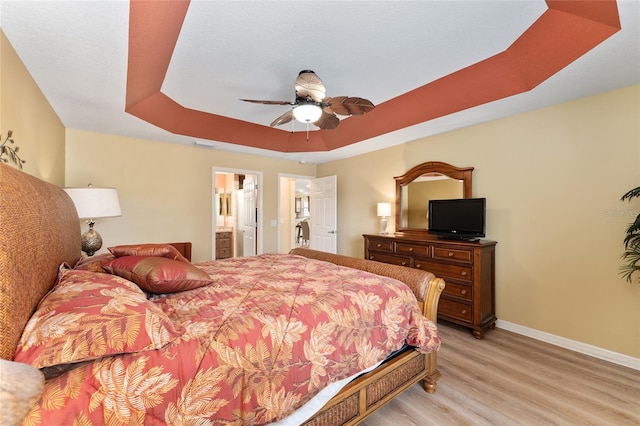 bedroom with baseboards, a ceiling fan, light wood-style flooring, ensuite bathroom, and a tray ceiling