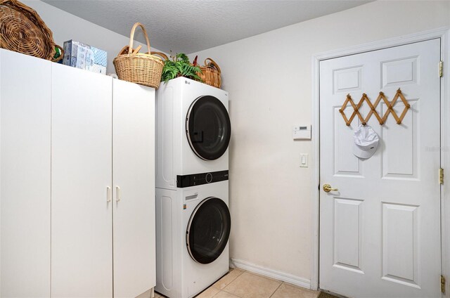 clothes washing area with light tile patterned floors, a textured ceiling, baseboards, stacked washing maching and dryer, and cabinet space