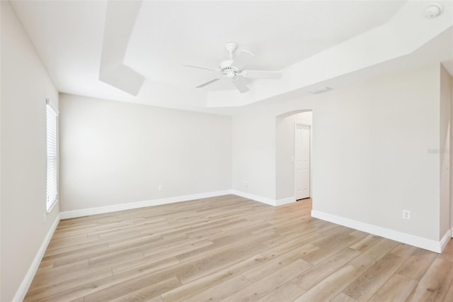 unfurnished room featuring a tray ceiling, arched walkways, light wood-style flooring, a ceiling fan, and baseboards