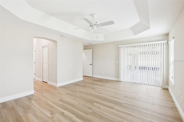 empty room featuring arched walkways, a tray ceiling, baseboards, and light wood-style floors