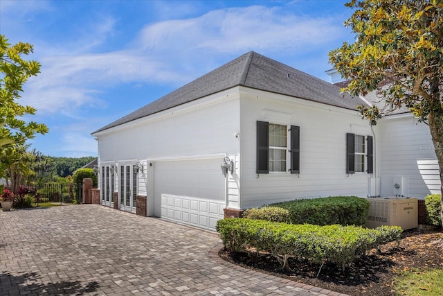 view of side of property featuring a garage, roof with shingles, fence, decorative driveway, and central air condition unit