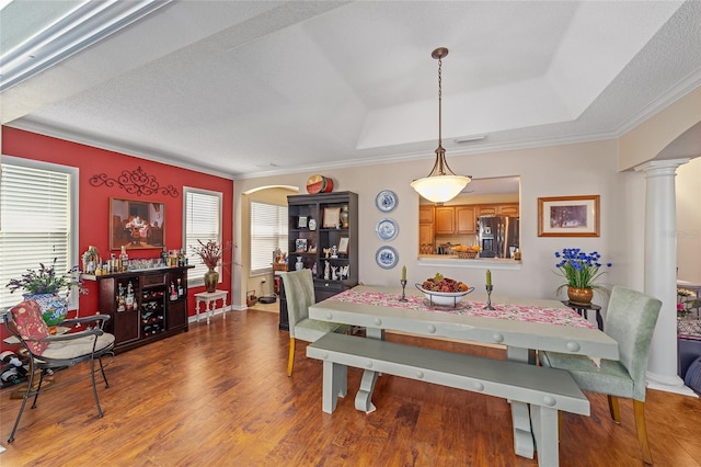 dining space with arched walkways, wood finished floors, ornamental molding, a tray ceiling, and decorative columns