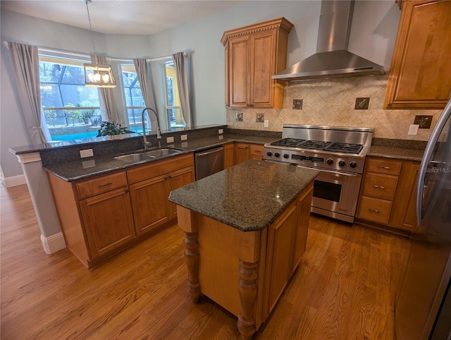 kitchen with a peninsula, stainless steel appliances, a sink, wall chimney exhaust hood, and brown cabinetry