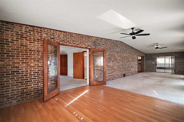 unfurnished living room featuring lofted ceiling with skylight, light wood finished floors, brick wall, and a ceiling fan