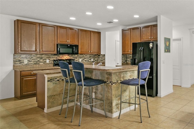 kitchen featuring visible vents, an island with sink, brown cabinets, black appliances, and a kitchen bar